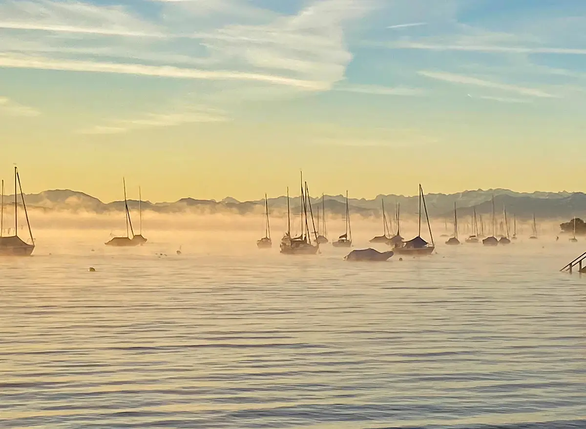 Das Südufer des Ammersees bei Sonnenaufgang mit Booten im Nebel und Bergkulisse